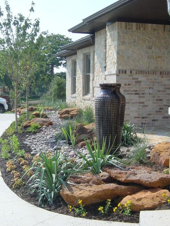 Modern landscape curbing features decorative concrete edging with a tall vase, surrounded by pebbles and ornamental grasses, enhancing curb appeal.