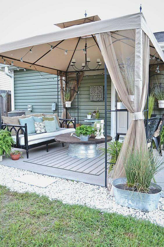 Outdoor patio with a steel canopy and tan fabric, featuring a sectional sofa, round coffee table, potted plants, and decorative rug.