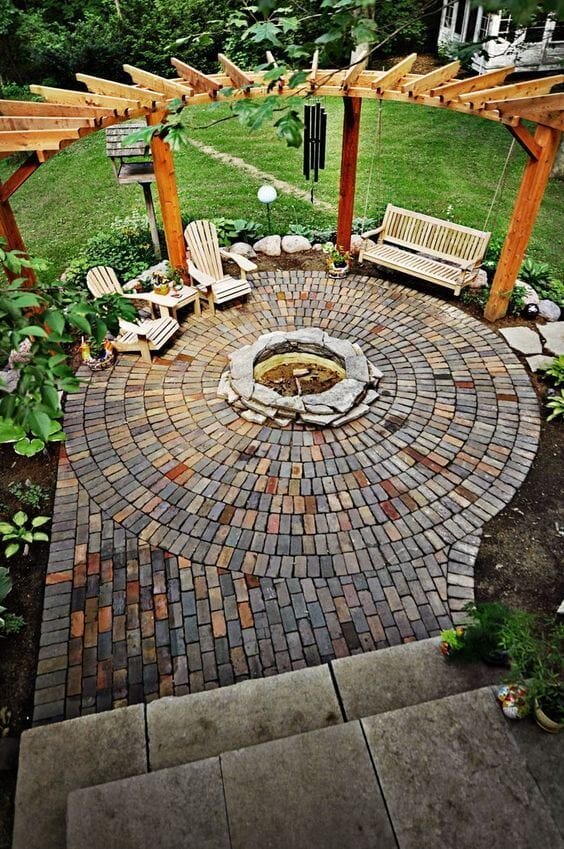 Circular patio with interlocking brick pavers features a natural stone fire pit, Adirondack chairs, and a bench swing under a pergola.