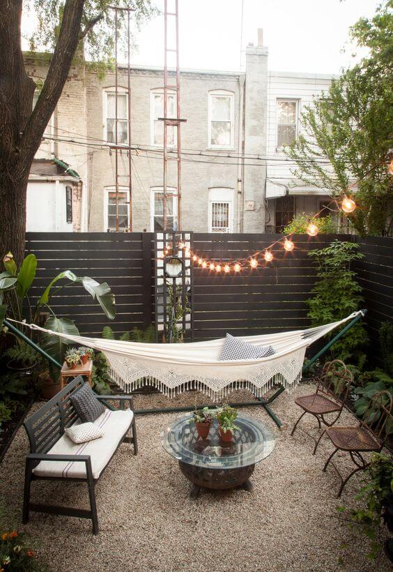 Outdoor living space with a hammock, gravel surface, surrounding plants, circular glass table, wrought iron chairs, and string lights for ambiance.