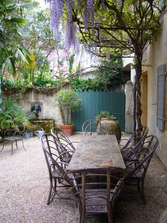 A stone rectangular table with wooden support and eight metal-framed chairs is set in a compact courtyard, surrounded by lush greenery.