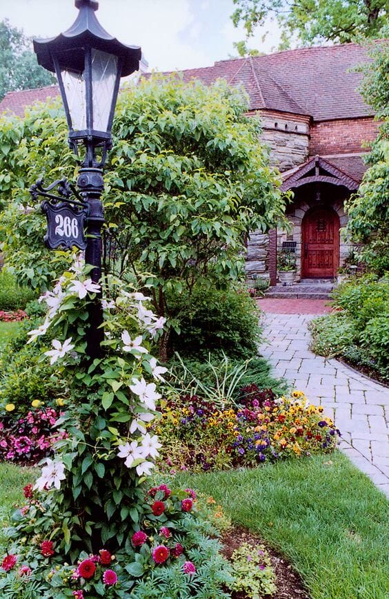 Elegant flower landscape design featuring a pathway, vibrant flowering plants, and a lamp post adorned with climbing blossoms. Lush green grass and colorful annuals create a welcoming atmosphere.