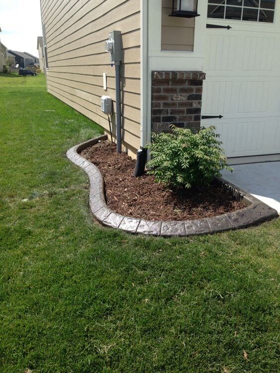 Curved garden bed with polished mulch and a small shrub, bordered by concrete landscape curbing, enhancing drainage and aesthetics.