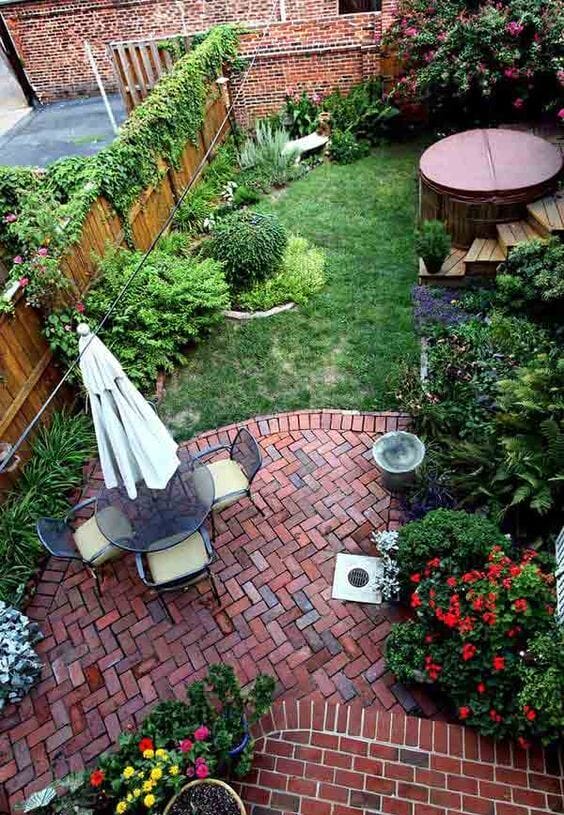 Backyard garden design with a brick patio, circular seating, glass table, and diverse plantings, enhancing relaxation in a compact space.
