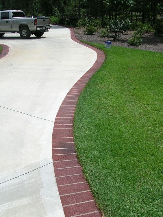 Curved concrete driveway edged with red brick pavers, surrounded by a manicured lawn and evenly spaced shrubs, enhancing landscape aesthetics.