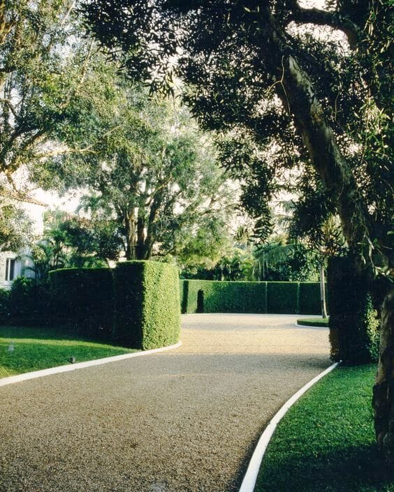 Curved concrete curbing outlines a gravel driveway, bordered by trimmed hedges and lush trees, enhancing the landscape design.