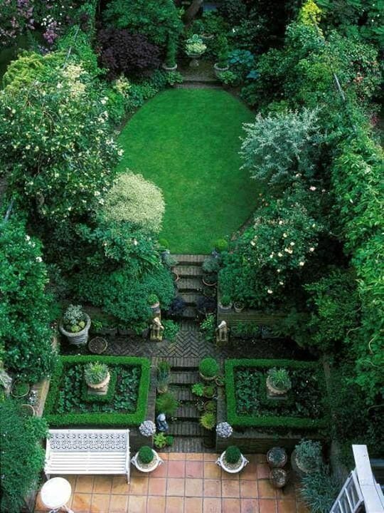 Central oval lawn surrounded by flower beds and greenery, with herringbone brick pathways, merging formal and informal garden styles.