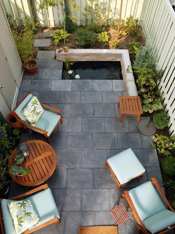 Compact backyard landscaping features a rectangular pond with stone edging, surrounded by slate tiles, wooden lounge chairs, and ornamental plants.