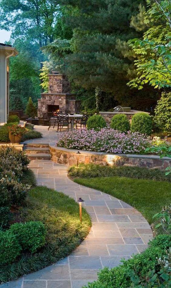 Winding stone pathway leads through a vibrant flower landscape with rounded pink beds, manicured boxwood, and a stone fire pit, framed by conifers.