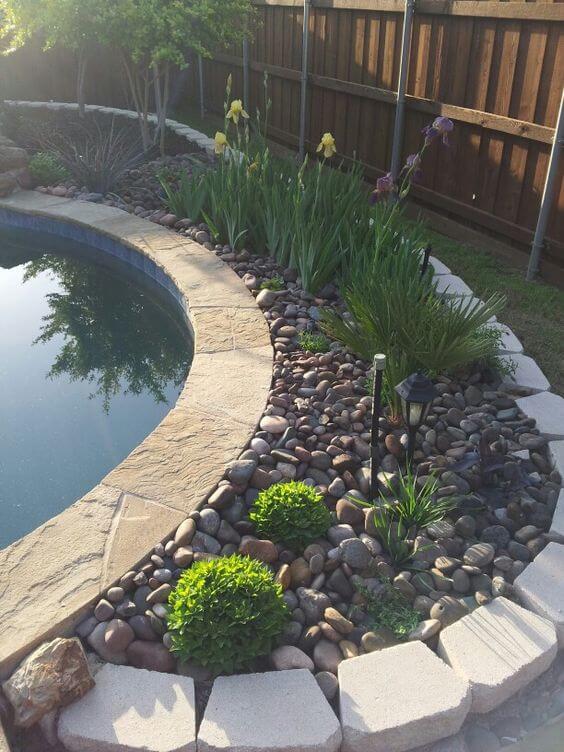 Curved landscape curbing features natural stone coping around a pool, with river rocks, low-maintenance shrubs, and flowering plants.