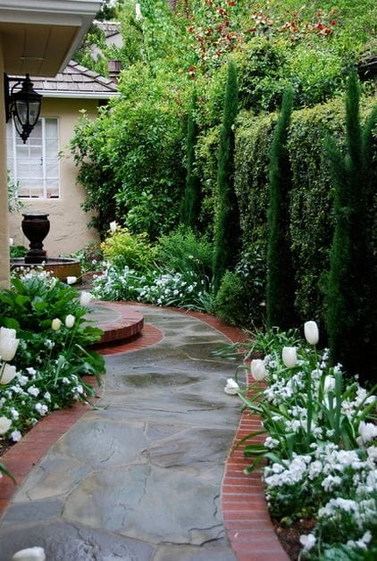A winding slate stone pathway, bordered by red bricks, flanked by evergreen shrubs and clusters of white tulips, creates a vibrant flower landscape.