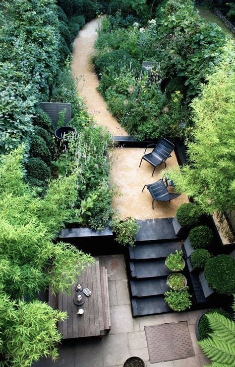 Secluded garden with a winding gravel path, bordered by evergreen shrubs. Features modern black chairs on a slate platform and a wooden table.