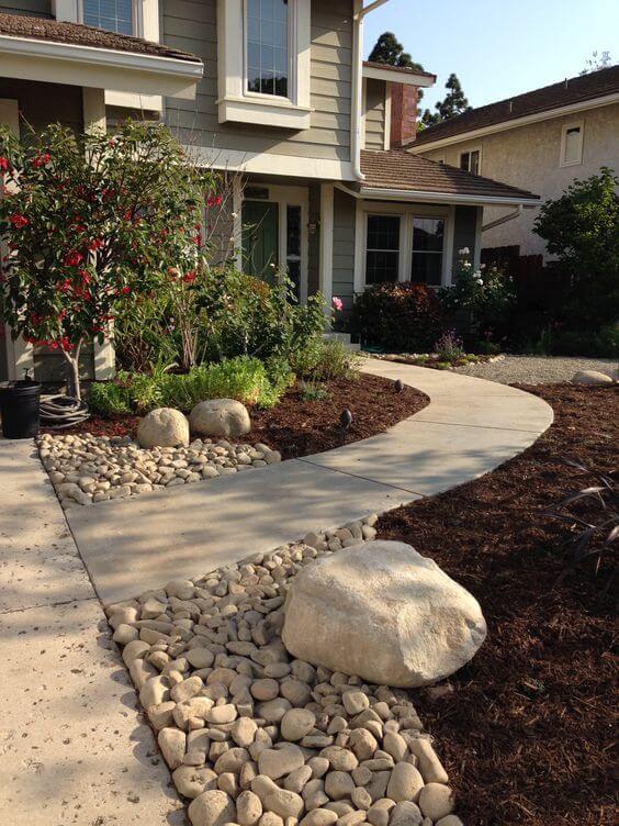 Curved concrete walkway connects entrance to driveway, bordered by river rocks. Lush greenery and mulch enhance a budget-friendly backyard design.