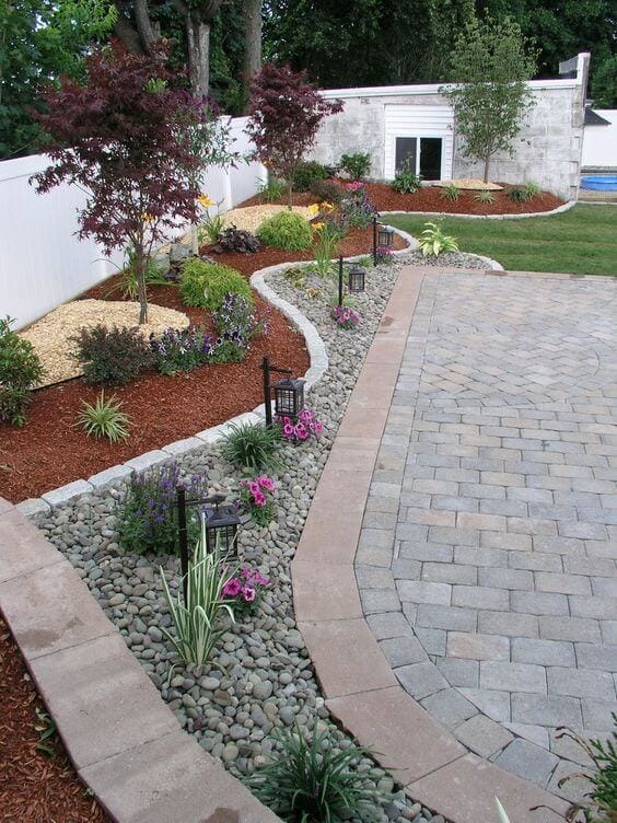 Curved stone pathway in a landscaped garden, bordered by colorful flower beds, featuring river rocks and perennial plants for visual interest.