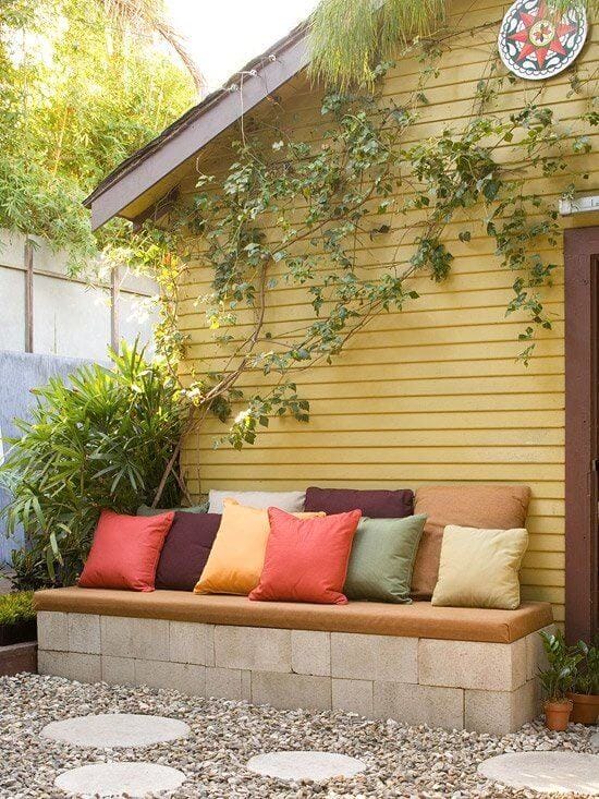 Outdoor seating area with a concrete block bench, cushioned seats, colorful pillows, gravel ground, and lush greenery, ideal for budget landscaping.