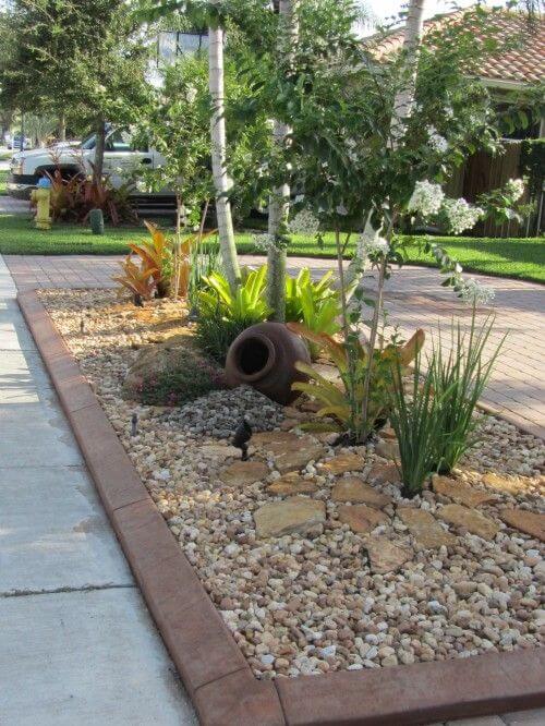 A landscaped bed with concrete curbing, featuring tropical plants, decorative stones, and a terracotta pot, enhancing visual interest.