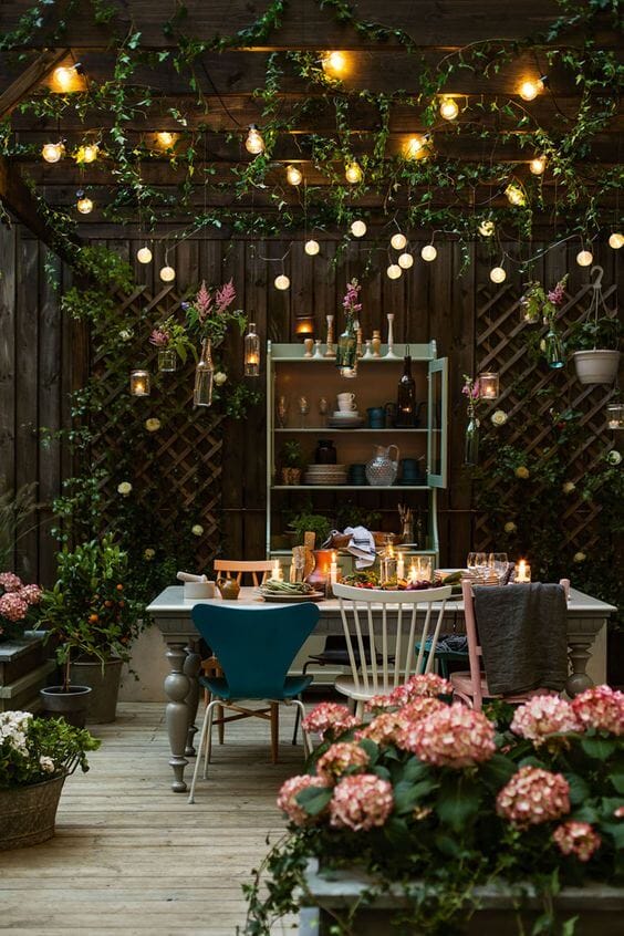 Backyard landscaping features a cozy outdoor dining space with a wooden deck, pastel chairs, climbing plants, and warm lighting, ideal for gatherings.
