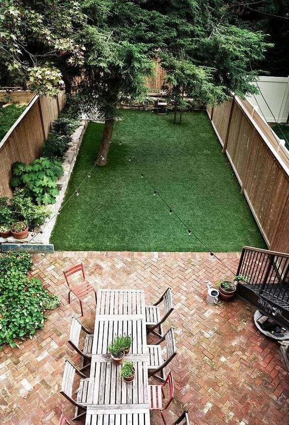 Backyard features synthetic grass, a herringbone brick patio with a wooden dining table for eight, and string lights for ambiance.