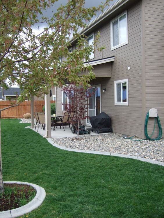 Landscaped area with well-maintained grass, curved cement pathway, and seating under an awning, surrounded by flowering trees and shrubs.