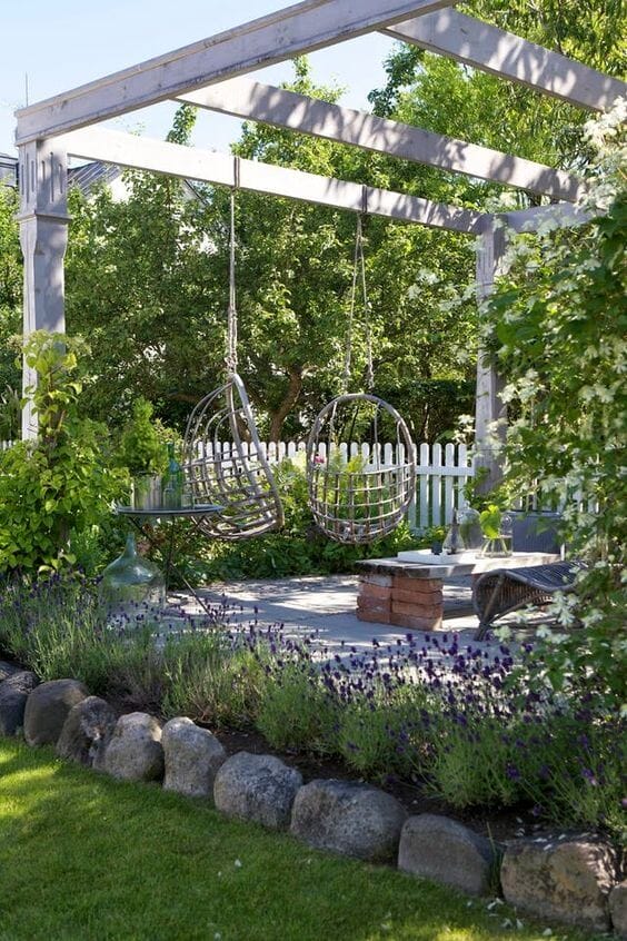 Hanging chairs under a wooden pergola, surrounded by lavender plants, create a cozy, rustic outdoor space with a stone-paved patio.