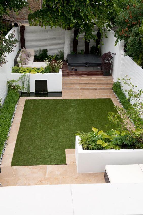 Modern backyard landscaping features a central synthetic grass area, surrounded by low-maintenance shrubs and concrete pavers. A cozy seating area and hot tub enhance relaxation.