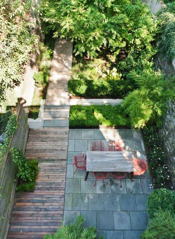 Compact urban garden with a stone patio, lush greenery, and a wooden walkway. Features a wooden table and red chairs for a vibrant, relaxing space.