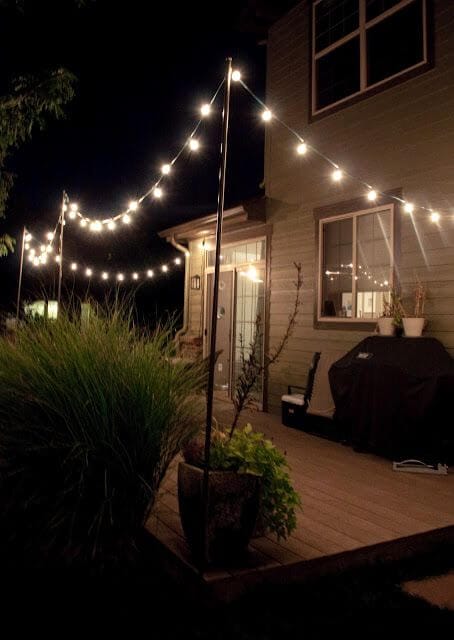 Overhead string lights suspended between poles illuminate a patio, enhancing the ambiance for evening gatherings and highlighting lush landscape features.