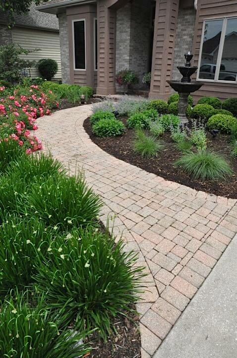 Curved interlocking paver path leads to a front entrance, flanked by vibrant pink azaleas and lush ornamental grasses, enhancing landscape design.