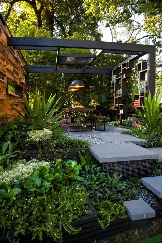 Structured outdoor seating area under a black metal canopy, featuring a wooden table and diverse plantings in tiered raised beds.
