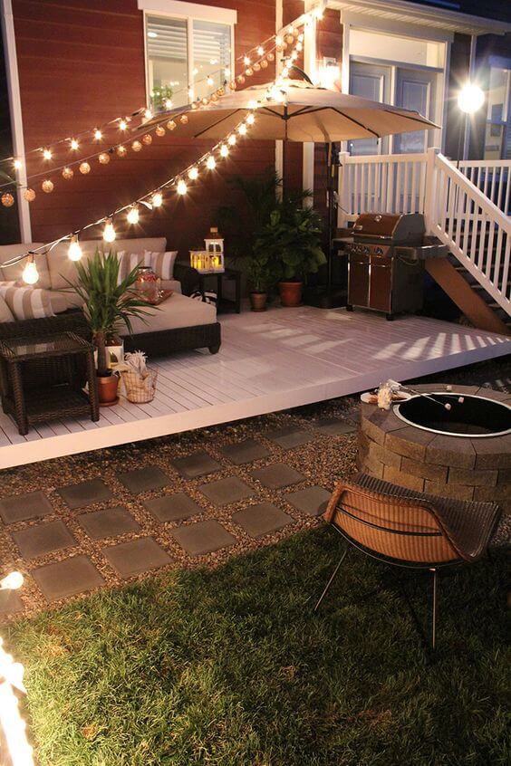 Backyard patio with a wooden deck, fire pit, dark wicker sofa, and ambient lighting, surrounded by stone pavers and plants.