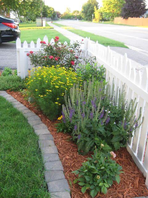 Curved stone border delineates a vibrant flower bed with yellow coreopsis, red roses, and purple salvia, enhancing front yard landscaping.