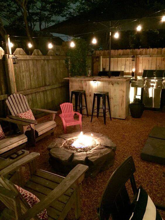 Cozy backyard fire pit design featuring a circular stone fire pit, wooden seating, vibrant pink children's chair, and ambient hanging lights.