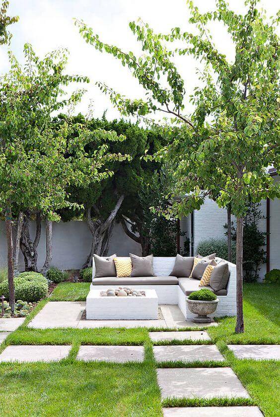 Outdoor seating area with a modern sectional sofa, stone pavers on a green lawn, and trees providing shade for a cozy backyard fire pit.