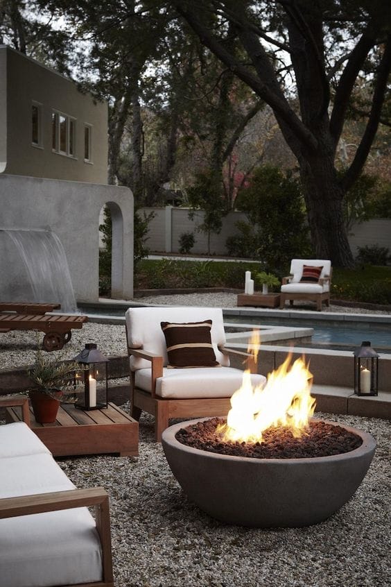 Cozy outdoor lounge features a circular concrete fire pit with lava rocks, two upholstered chairs, and a serene water feature backdrop.