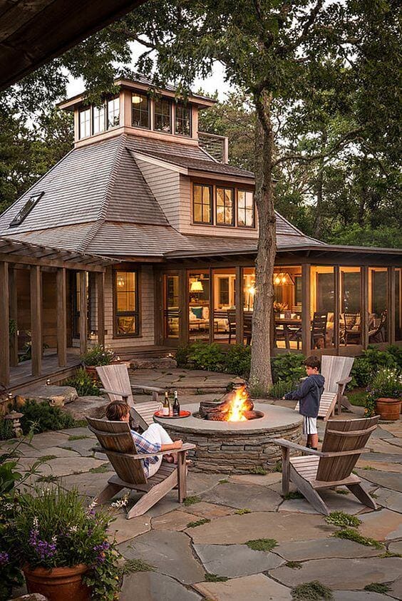 Circular stone fire pit surrounded by natural slate pavers, featuring Adirondack chairs and lush greenery, ideal for cozy gatherings.