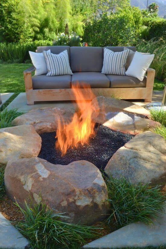 Circular fire pit made of large boulders, filled with black lava rock. A contemporary gray sofa with striped cushions is nearby, surrounded by lush greenery.