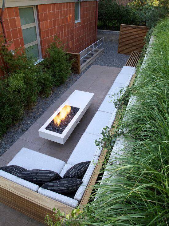 Outdoor lounge with a built-in fire pit as the focal point, featuring L-shaped modern benches, concrete paver flooring, and surrounding greenery.