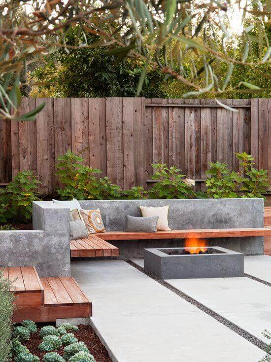 Modern backyard fire pit design featuring an L-shaped concrete bench, dark concrete fire pit, and lush greenery for a cozy ambiance.