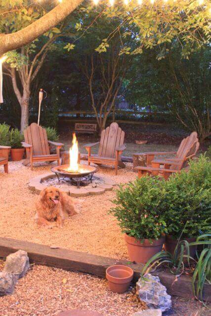 Cozy backyard fire pit area with a central stone fire pit, four Adirondack chairs, string lights, and potted plants for ambiance.