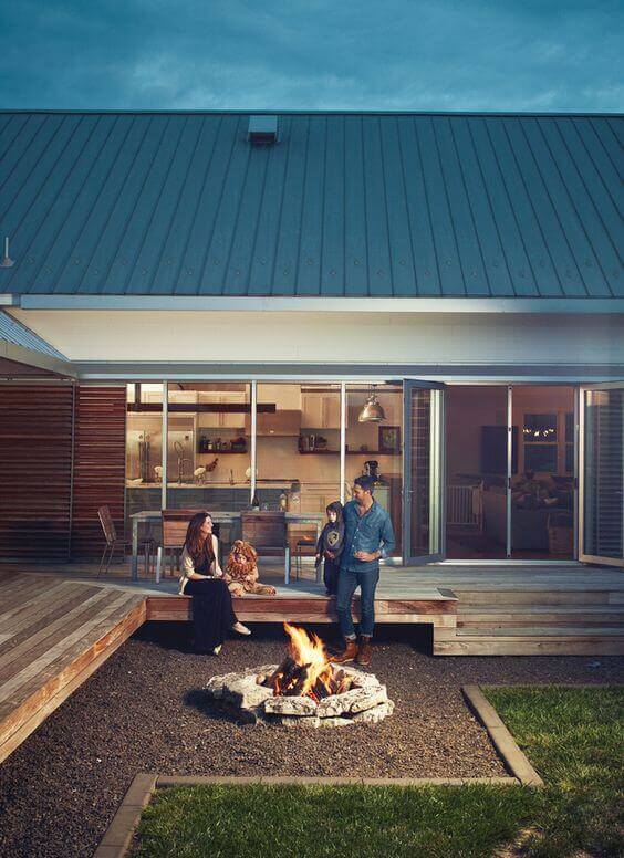 Circular stone fire pit at the center of a gravel landscape, bordered by a wooden deck, featuring large glass doors for indoor-outdoor flow.