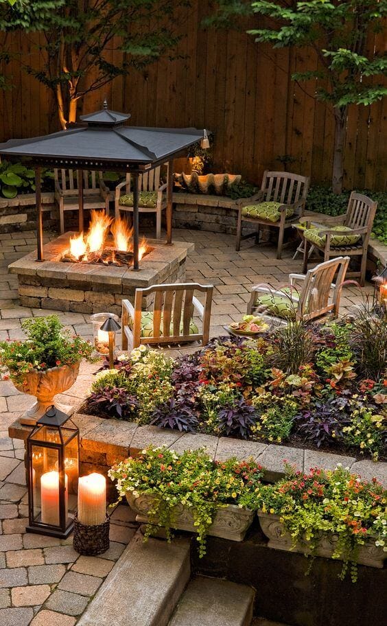 Outdoor fire pit area with a central stone fire pit, six wooden chairs, interlocking paver circle, gazebo, and ambient lighting for cozy gatherings.