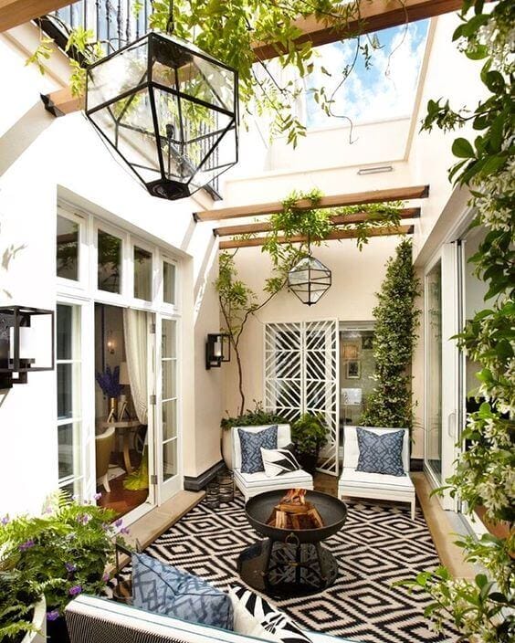 Modern patio design featuring a black metal fire pit surrounded by white lounge chairs and a patterned rug, enclosed by light walls and greenery.