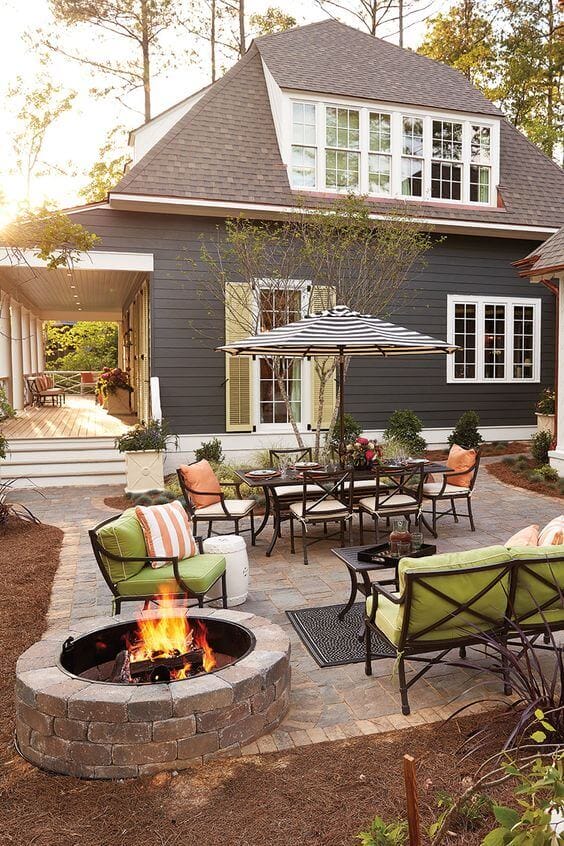 Outdoor patio with a circular stone fire pit as the centerpiece, surrounded by a retaining wall, dining table, and lounge chairs.
