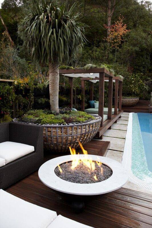Modern backyard fire pit design features a round concrete fire pit surrounded by gravel, with natural stone pavers and wooden decking.