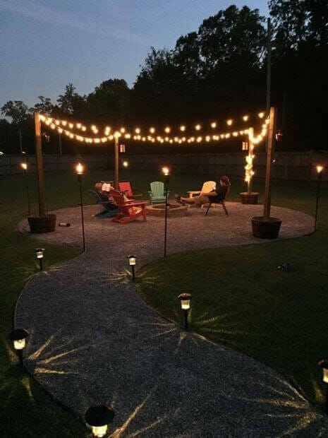 Cozy backyard fire pit area with a circular gravel surface, colorful chairs, and string lights for ambiance, surrounded by grassy space.