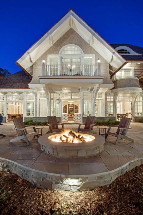 Circular stone fire pit on a flagstone patio, surrounded by six Adirondack chairs, creating a cozy gathering space near a two-story home. Soft lighting enhances the evening ambiance.