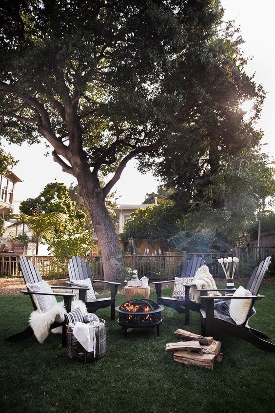 Cozy outdoor seating area with five dark wood Adirondack chairs around a 36-inch metal fire pit, surrounded by well-kept grass and a mature tree.