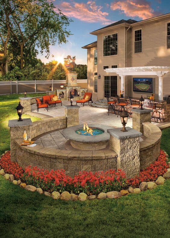 Circular patio with interlocking stone pavers, central gas fire pit, low seating walls, vibrant red floral borders, and shaded dining area under a pergola.
