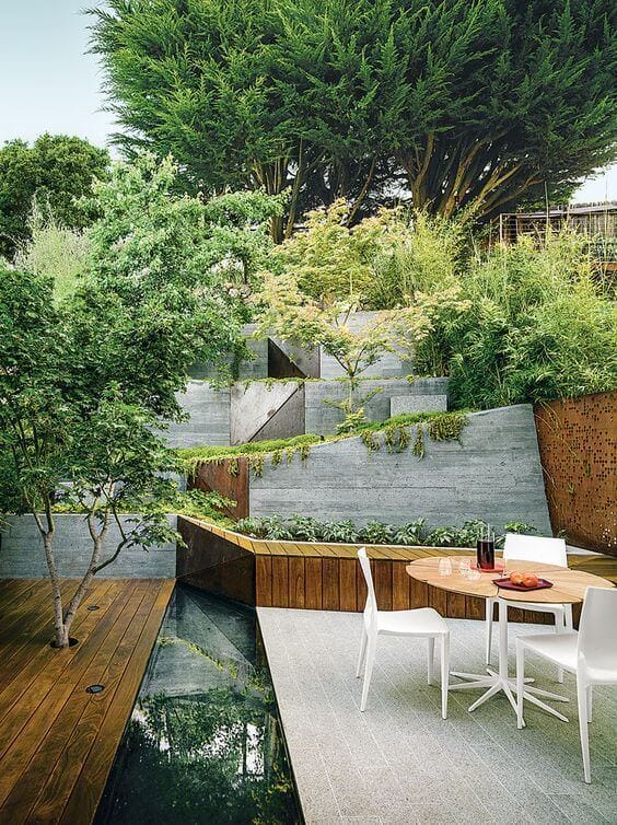 Tiered garden with concrete retaining walls, tropical plants, a wooden deck, and a circular dining set, designed for sustainability and aesthetics.