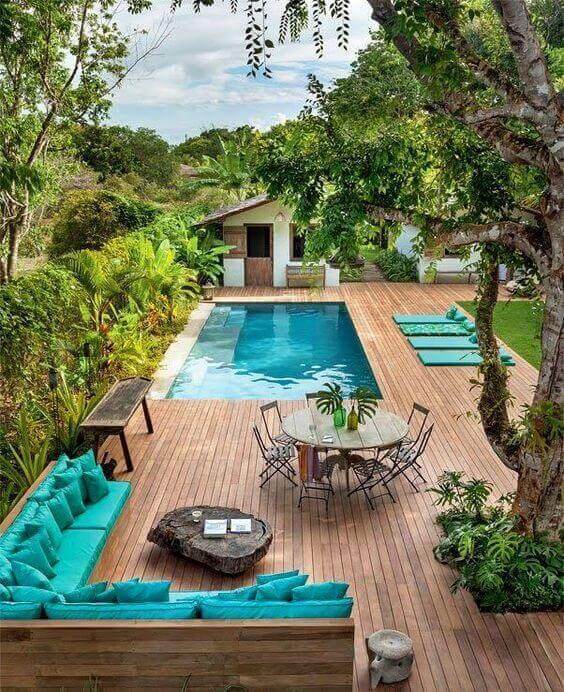 Rectangular swimming pool (5x10m) surrounded by hardwood deck. Features turquoise cushions, dining table, tropical plants, and lounging pads.